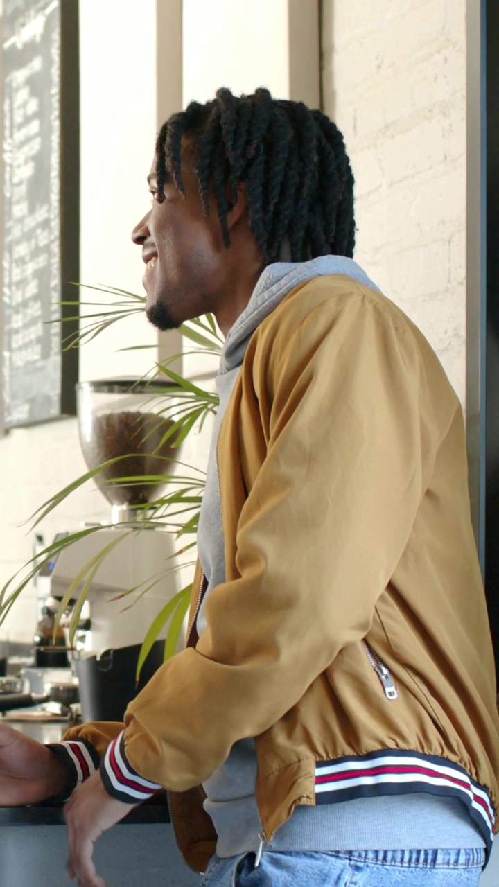 Barista che serve, uomo con giacca color cammello e felpa grigia con cappuccio che si appoggia e ordina al bancone del caffè consultando il menu.