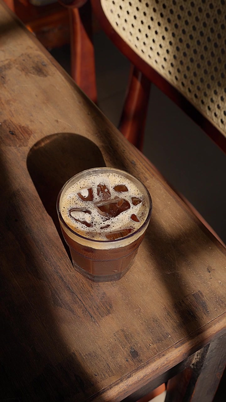 Primo piano di un rinfrescante caffè freddo marrone scuro su un tavolo di legno antico. La luce del sole dalla finestra proietta ombre suggestive sul vetro e sul legno texturizzato, creando un'atmosfera vintage nostalgica