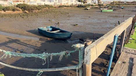 Spaziergang-über-Eine-Alte-Und-Verfallene-Holzpromenade-Mit-Ausgetrockneter-Und-Schlammiger-Bodenoberfläche,-Altem-Fischerboot-Und-Reifen-Am-Leeren-Flussgrund