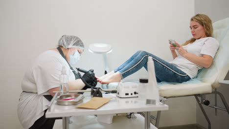 beautician in mask and gloves using electric nail filer on client lying on chair while client browses phone during pedicure session, with various manicure tools and products placed on white table