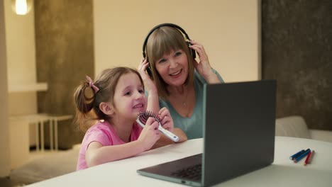 Kleines-Mädchen-Sitzt-Am-Schreibtisch-Am-Laptop-Und-Singt,-Während-Großmutter-In-Der-Nähe-Musik-über-Kopfhörer-Hört