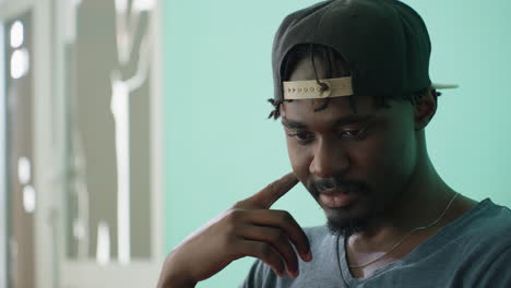 close up of calm young man wearing cap looking down thoughtfully indoors with soft light and blurred background, creating reflective and focused atmosphere with artistic silhouette shape on wall