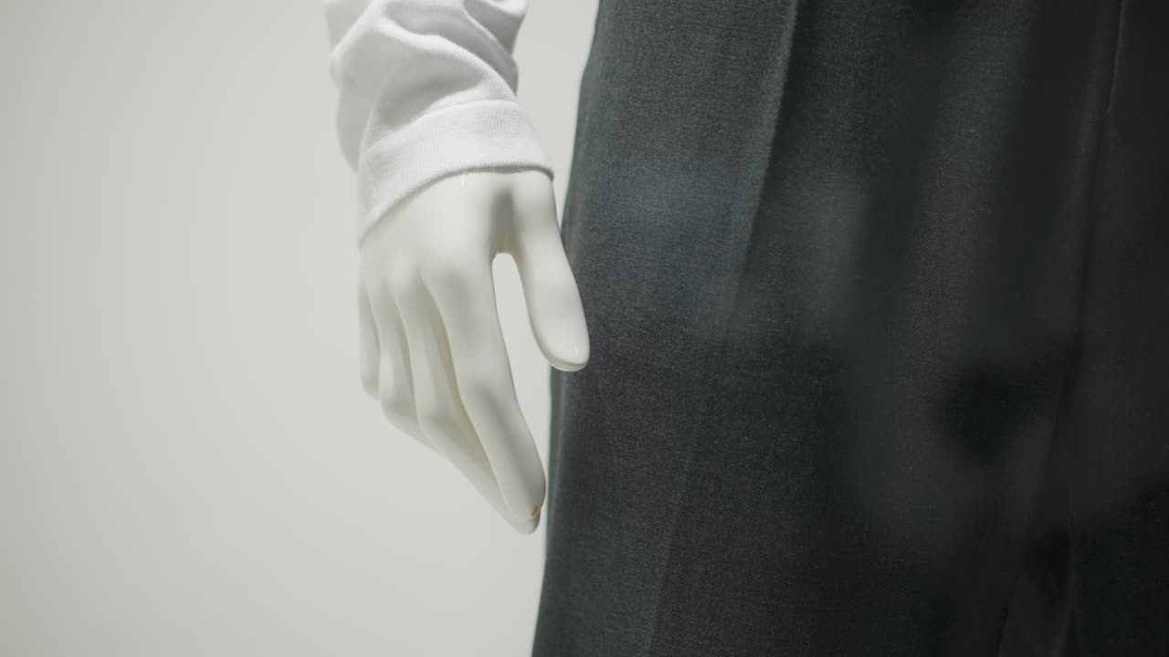 vista parcial de la mano del maniquí con manga blanca larga y pantalones negros, mostrando detalles elegantes de la ropa con iluminación suave, composición minimalista y fondo limpio