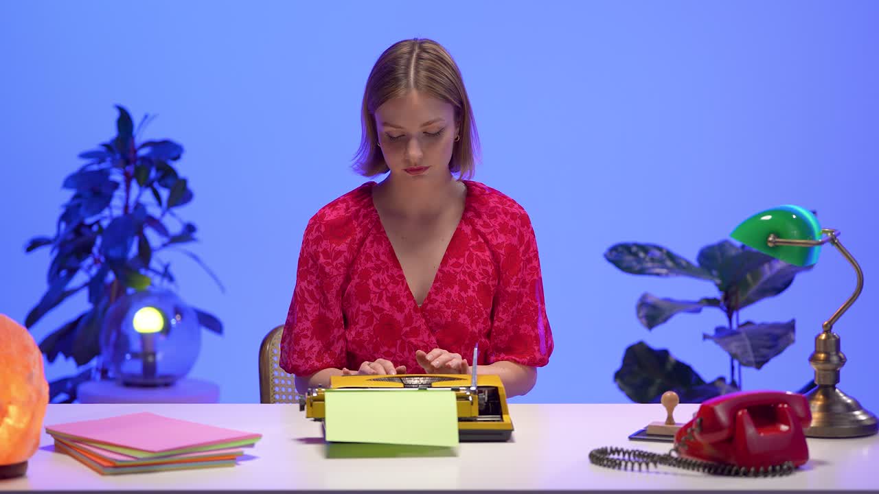 A woman in a red floral dress types attentively on a yellow typewriter at a retro-inspired desk. Surrounded by nostalgic props and pastel accents, she works in a creative and colorful setting