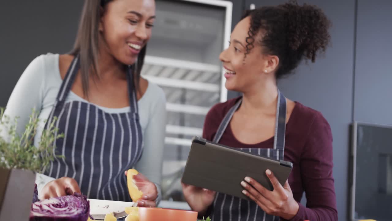 feliz pareja de lesbianas biraciales en delantales preparando comida y usando tableta en la cocina, en cámara lenta