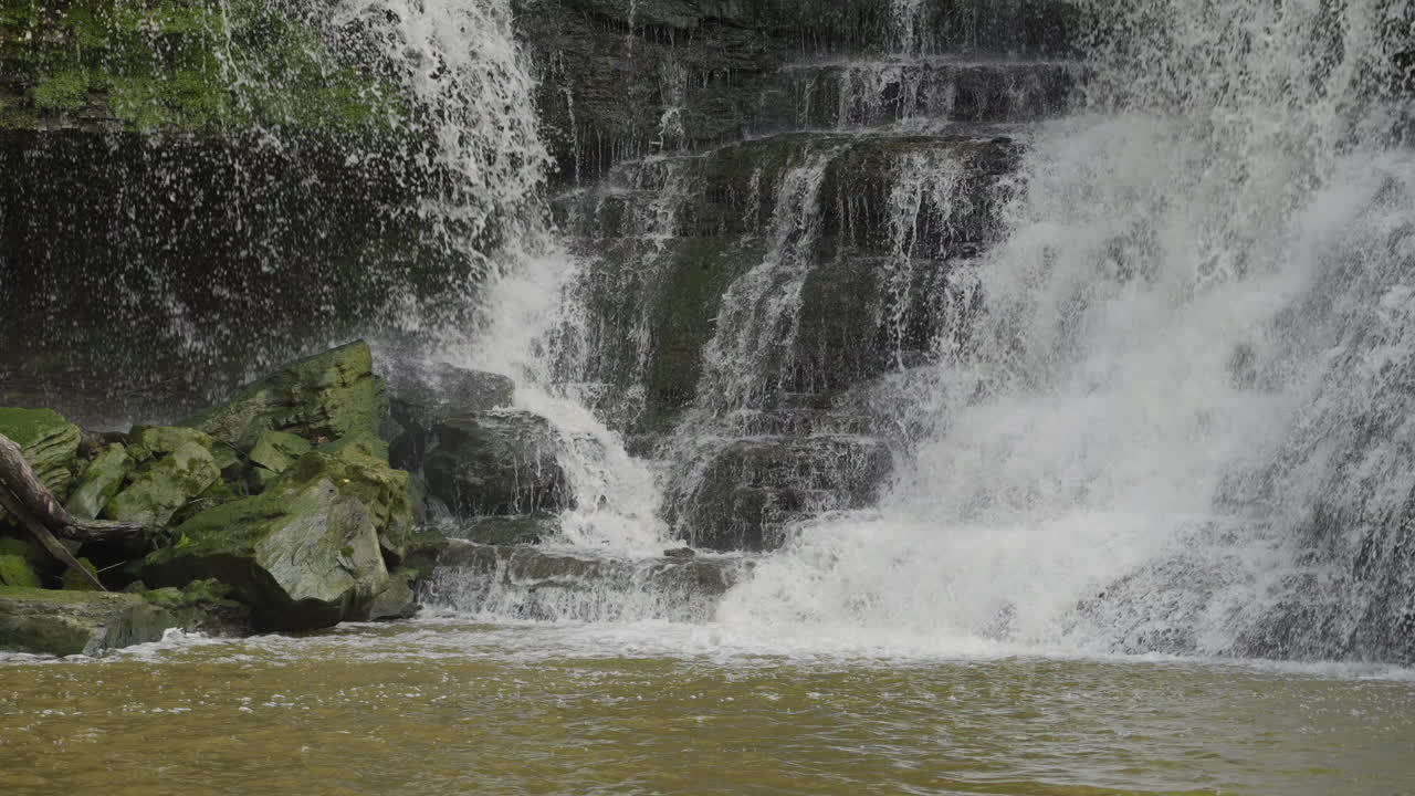 Slow motion close-up of waterfall splashing over green rocks on a sunny day