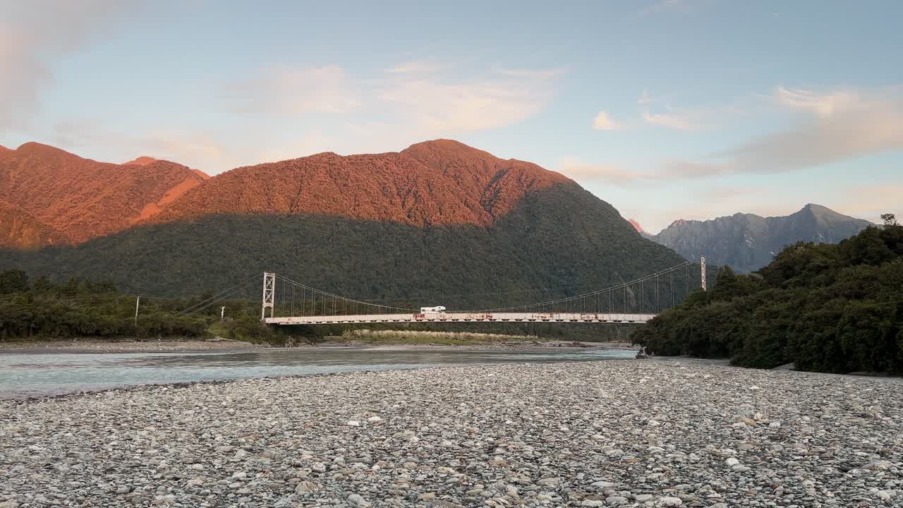 야영객은 해가 지는 동안 뉴질랜드의 외 ⁇  서부 해안 야생에서 오래된 녹색 강철 다리를 건너고 있습니다.