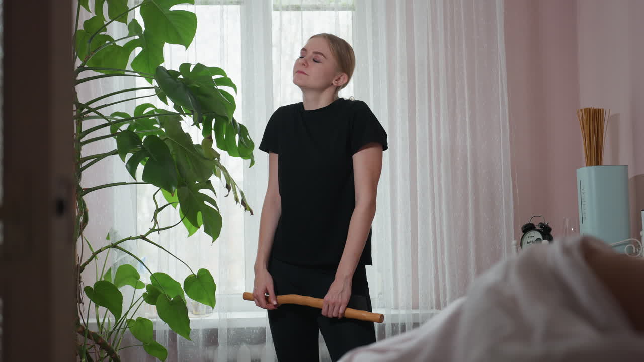 Tired woman in black outfit standing near large indoor plant holding wooden massage tool, eyes closed and head tilted back in exhaustion, set against bright window with sheer curtains