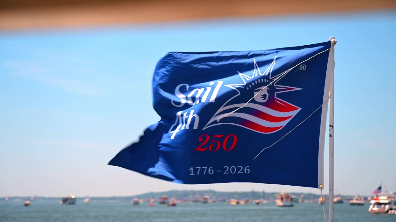 Sail4th 250-flag vajer over New York Harbor. Det blå banner fylder billedet mod en klar himmel, mens fjerne både samles på Uafhængighedsdagen