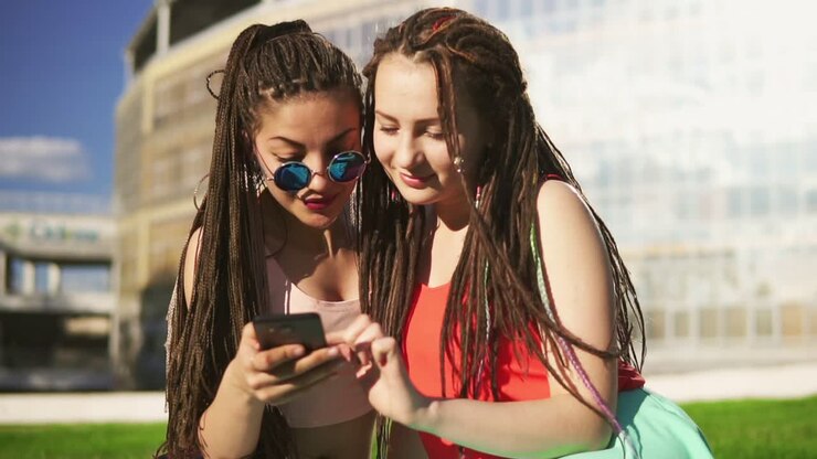 Happy women with dreads sitting on grass in summer park and using a smartphone. Young friends talking and watching photos