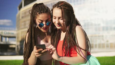 Glückliche-Frauen-Mit-Dreadlocks,-Die-Im-Sommerpark-Auf-Gras-Sitzen-Und-Ein-Smartphone-Benutzen.-Junge-Freunde-Reden-Und-Schauen-Sich-Fotos-An