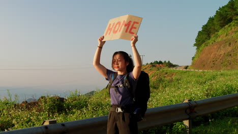 young solo female traveller hitchhiking on road side with a sign on cardboard saying home