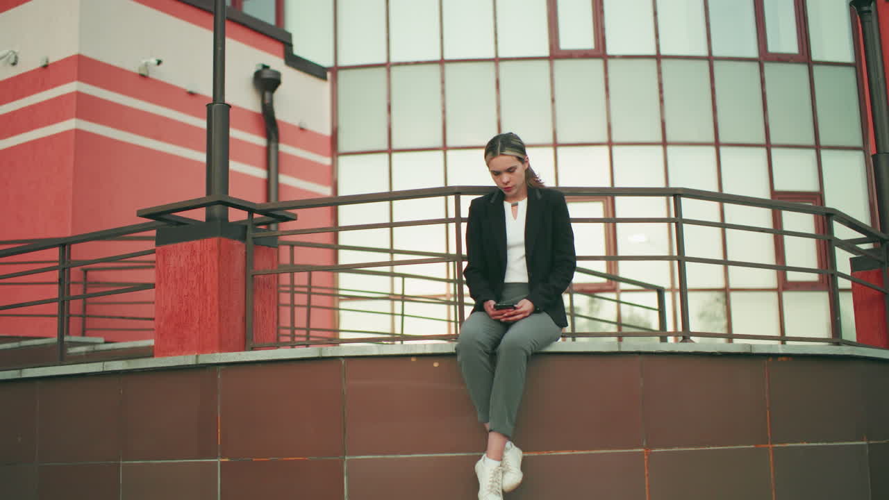 Young girl in black blazer seated on fence in urban area, holding phone while waiting for someone, legs crossed at ankle, modern glass building in background