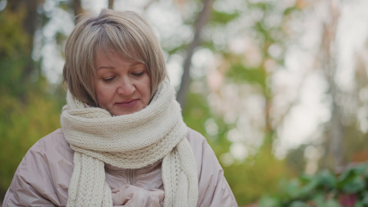 middle aged woman wearing thick cream scarf and pale jacket gazing downward with focused expression, background filled with blurred green and yellow trees in soft autumn forest ambiance