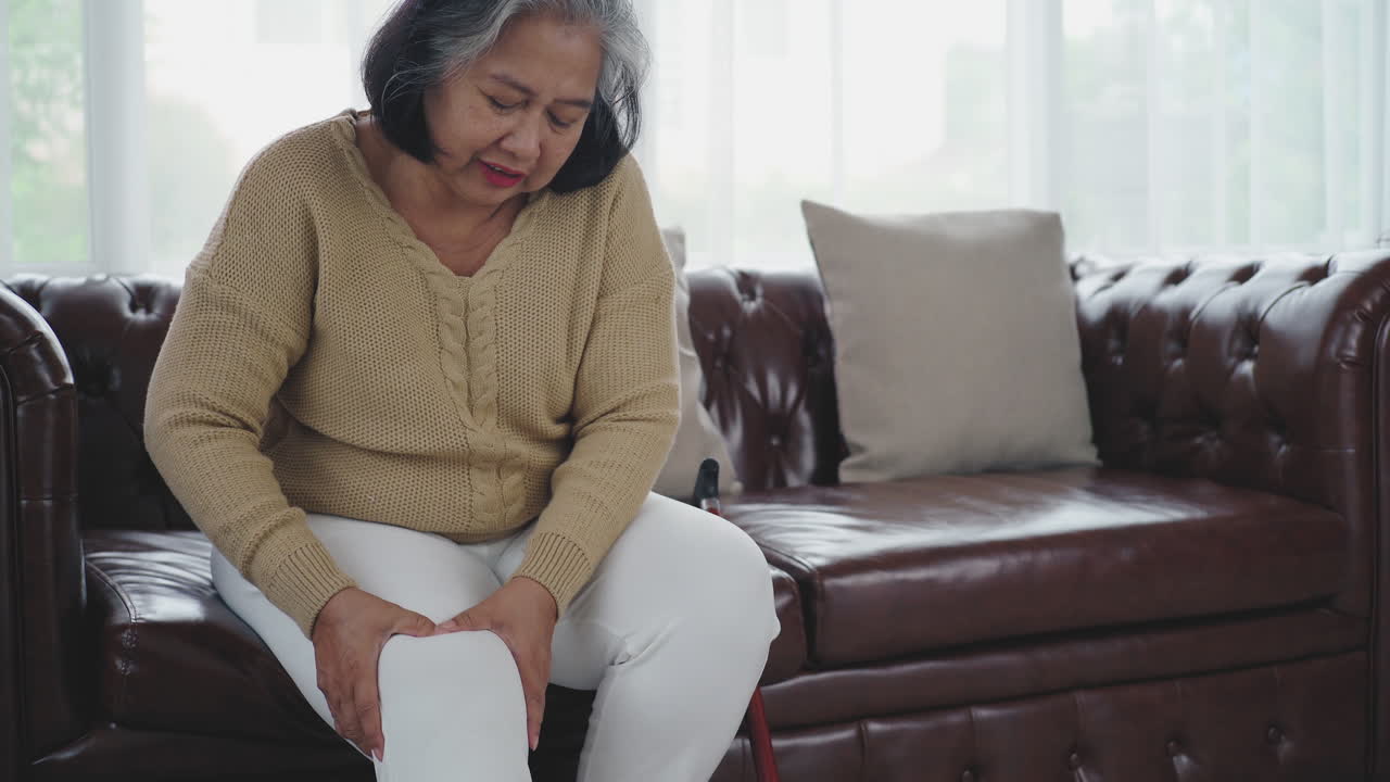 심각한 다리와 무릎 통증으로 고통스러운 표현을 하는 노인은 일어나서 소파에 앉아 집안을 돌아다니는 데 어려움을 겪습니다.