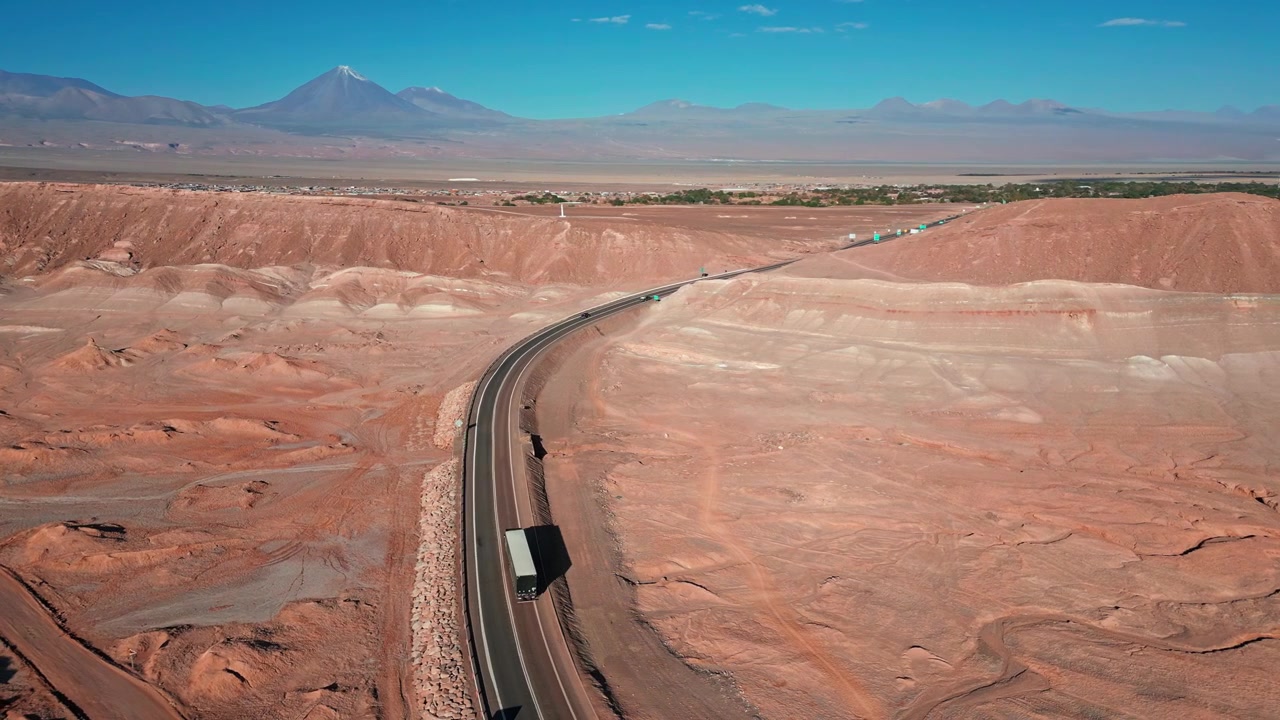 Panoramica aerea su autostrada tortuosa attraverso terreno desertico rosso con camion diretti verso l'oasi di San Pedro de Atacama e una cima vulcanica innevata oltre. San Pedro de Atacama, Cile