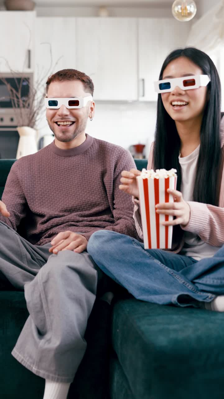 Pareja feliz viendo una película 3D y comiendo palomitas en el sofá.