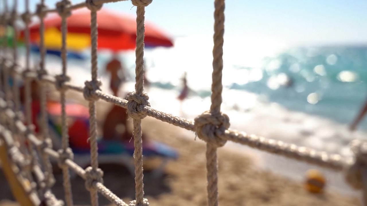 Una giornata serena in spiaggia immortalata attraverso una recinzione di corda annodata, che mostra ombrelloni vivaci, bagnanti e onde delicate sotto un cielo limpido.