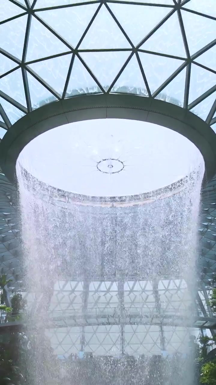 La cámara se inclina hacia arriba desde una gran cascada interior para revelar un techo de cúpula de cristal geométrico, con luz natural brillante iluminando el moderno espacio arquitectónico.