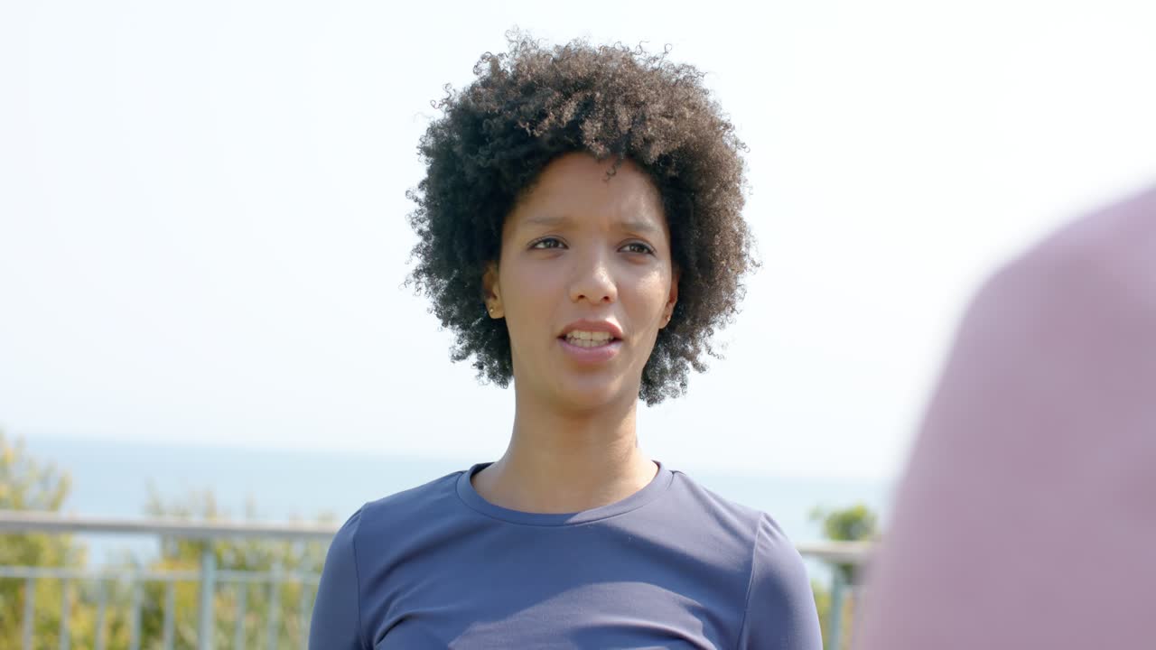 Mulher desviando o olhar para um gesto de mão fora da câmera e conversando em uma varanda à beira-mar.