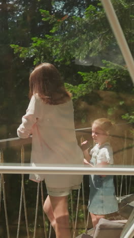 mother and daughter stand taking photos on glass balcony. little girl holds camera spending time against dense forest. happy family photograph against glamping park