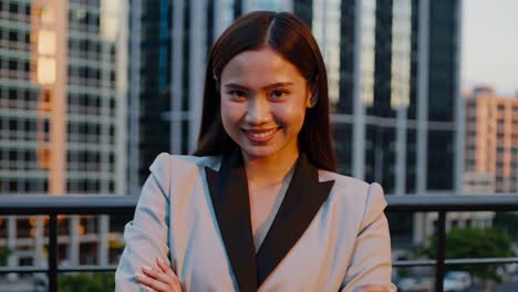 young asian businesswoman in elegant suit posing with crossed arms on rooftop with skyscrapers in background, looking at camera and smiling