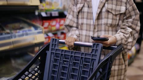 Unrecognizable-woman-with-mobile-device-and-trolley-purchasing-products-at-mall