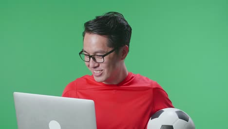 vista lateral de cerca de un hombre asiático con una pelota viendo un partido de fútbol en una computadora portátil en el estudio de pantalla verde