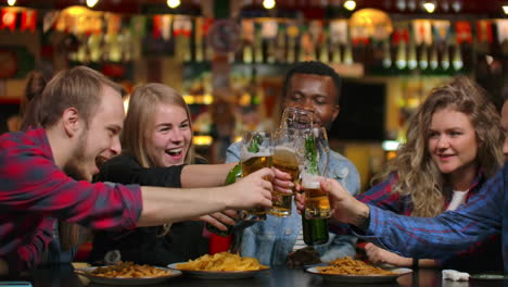 Plano-De-Cerca-Un-Grupo-De-Amigos-Hace-Un-Brindis-Y-Todos-Tocan,-Revisan-Sus-Vasos-Con-Cerveza,-Beben-Y-Ríen.-Estudiantes-Reunidos-En-Un-Bar-Restaurante.