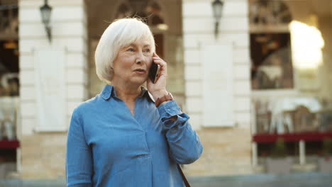 Grey-Haired-Old-Woman-Standing-In-City-And-Chatting-On-Smartphone