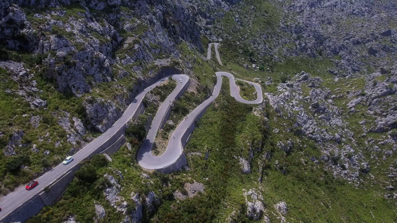 Vista aerea che si muove verso una strada di montagna a tornanti con veicoli che salgono e scendono attraverso un terreno roccioso a Maiorca