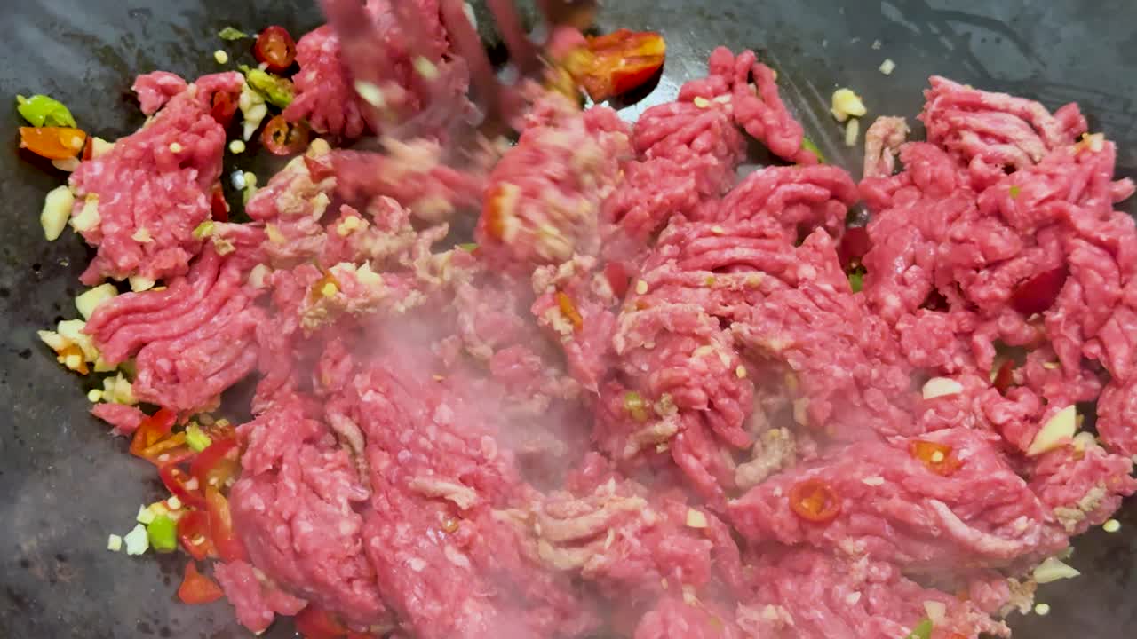 Raw minced meat is stir-fried with chopped vegetables and spices in a hot wok, using a spatula. Bright lighting, close-up, dynamic movement
