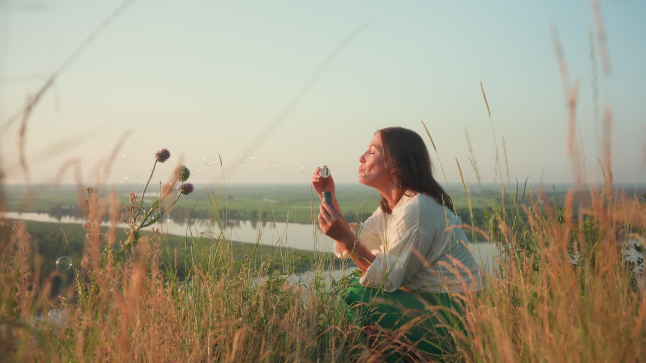 Sunset lit woman in green skirt squatting on grassy river bank blowing shimmering soap bubbles drifting over blurred valley of flower and river under warm golden light