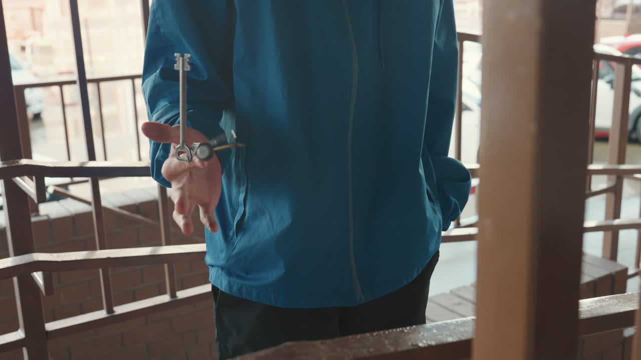 Person in blue jacket holding keys on wet balcony under drizzle, closeup of hand gripping metal railing, urban apartment backdrop, cinematic grain and moody lighting, subtle rain droplets on fabric