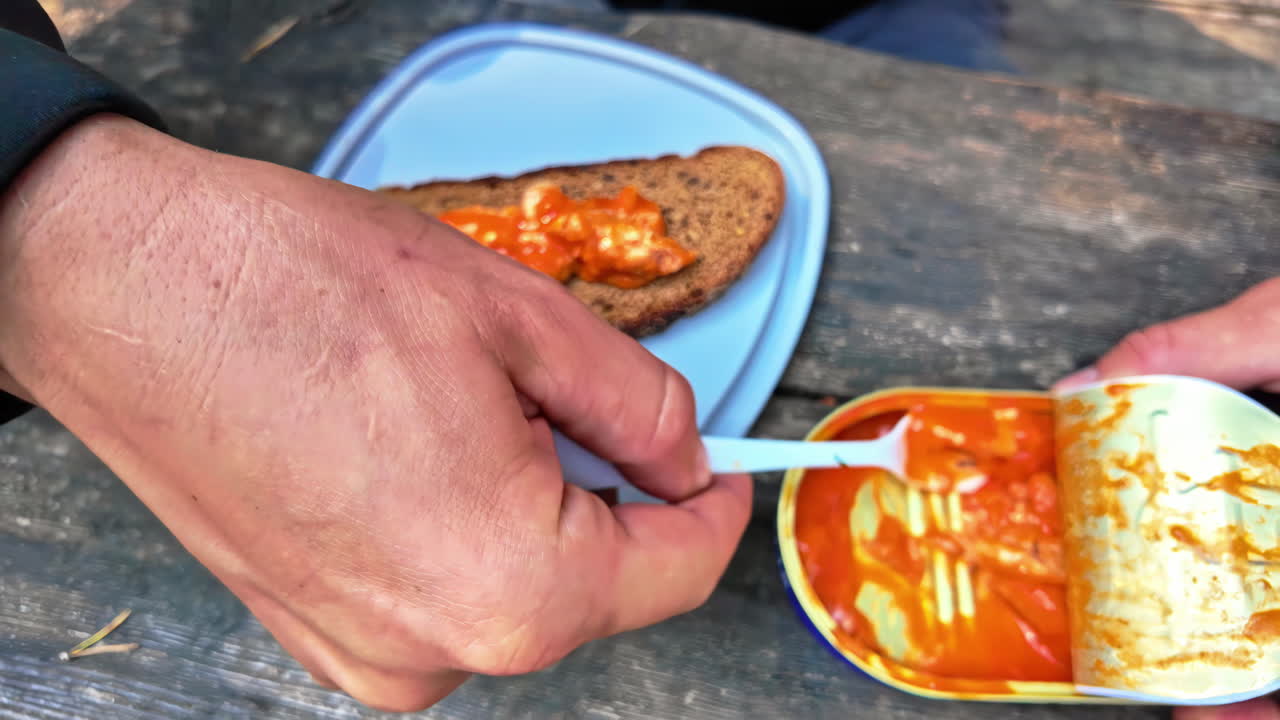 Closeup of person using plastic fork scooping sardines in sauce from can onto wooden picnic bench