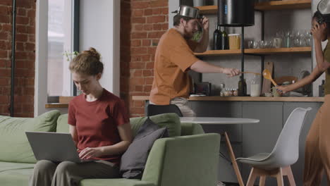 Chica-Trabajando-En-Una-Computadora-Portátil-Sentada-En-Un-Sofá-Mientras-Sus-Dos-Compañeros-De-Cuarto-Se-Divierten-Peleando-Con-Utensilios-De-Cocina-1