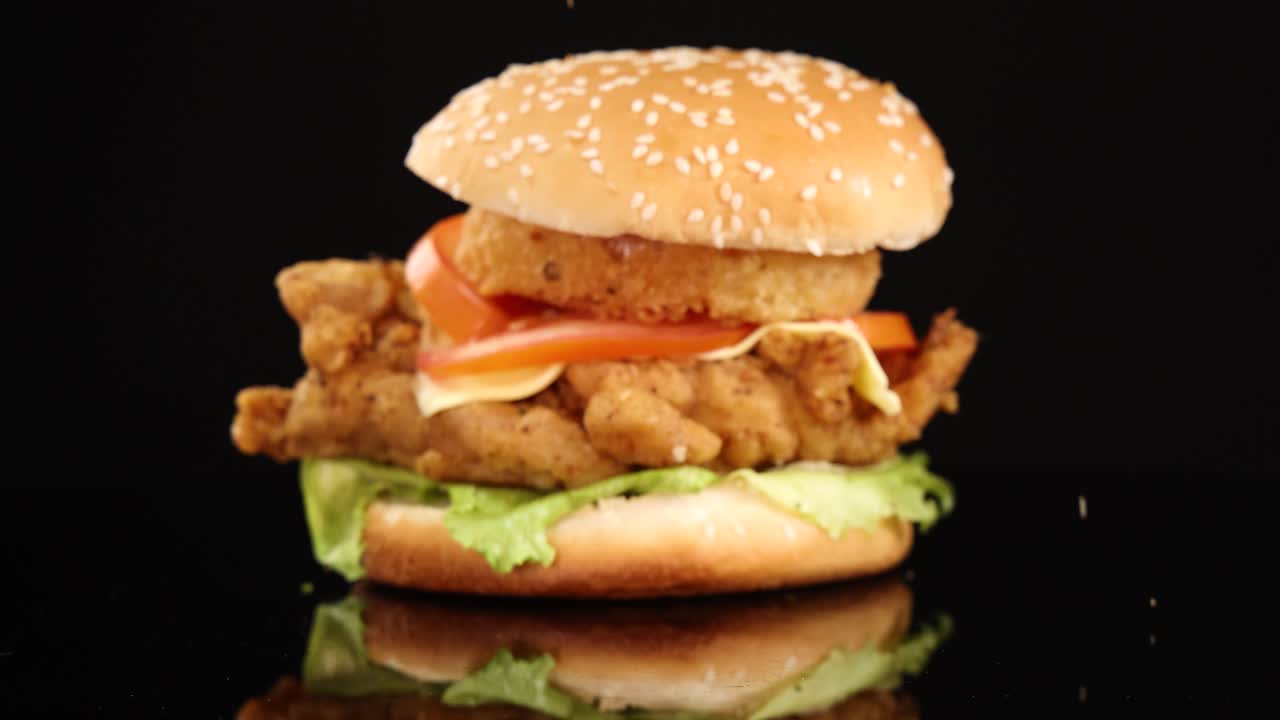 Golden onion rings fall in slow motion next to a fried chicken burger on a glossy black surface, under bright studio lighting with dramatic reflections