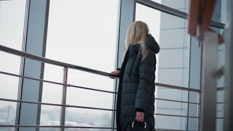 side view of woman in black jacket gazing through glass window, observing city traffic with cars moving in urban setting, creating a thoughtful and serene atmosphere in modern indoor space