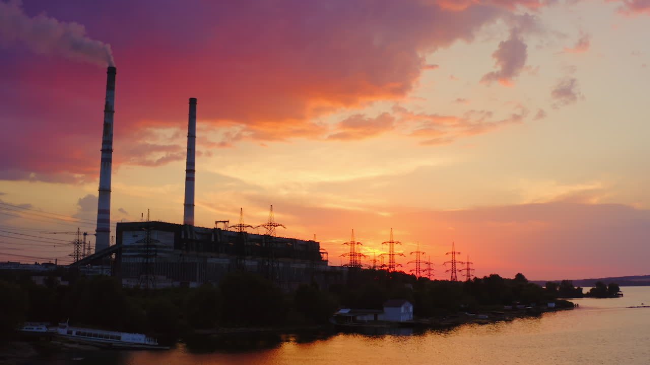 Industrial zone in the evening nature. Transmission lines, factory with pipe releases smoke near the river at sunset. View from the river.
