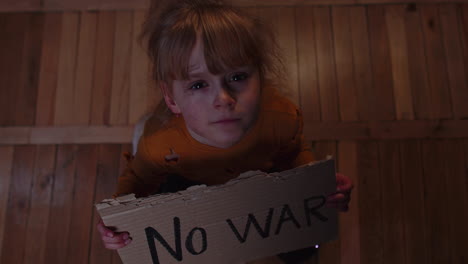 scared homeless toddler girl sitting holding inscription no war, hiding from bombing attack at home