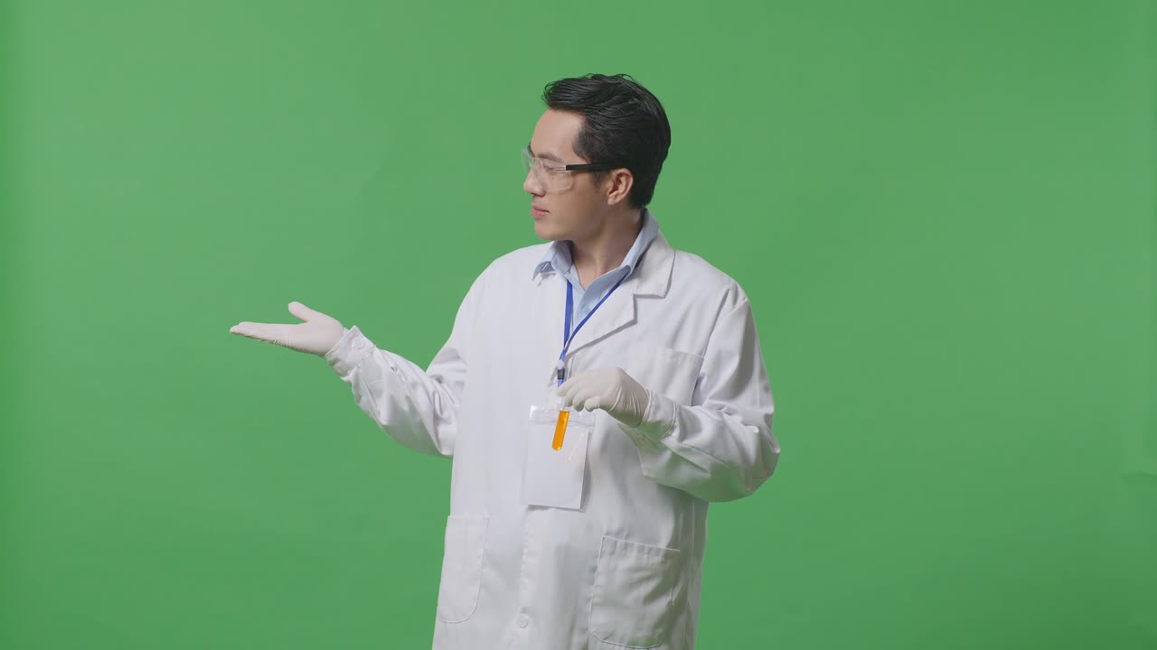실험실에서 녹색 화면 배경에 서 있는 동안 미소 짓고 으로 가리키는 실험 튜브에 오렌지 액체와 함께 아시아 남자 과학자