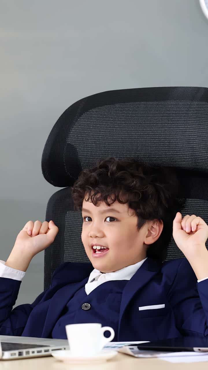 A young boy in a suit cheers and raises his arms while using a laptop
