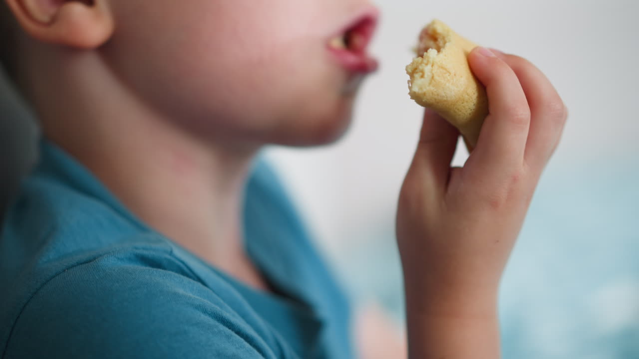 Close up of young white boy in blue shirt happily enjoying snack while holding cake with both hands against blurred vibrant background capturing pure childhood joy