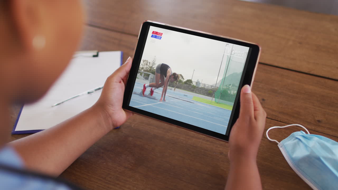 compuesto de una mujer sentada en una mesa con una máscara facial, viendo un evento de atletismo en la tableta