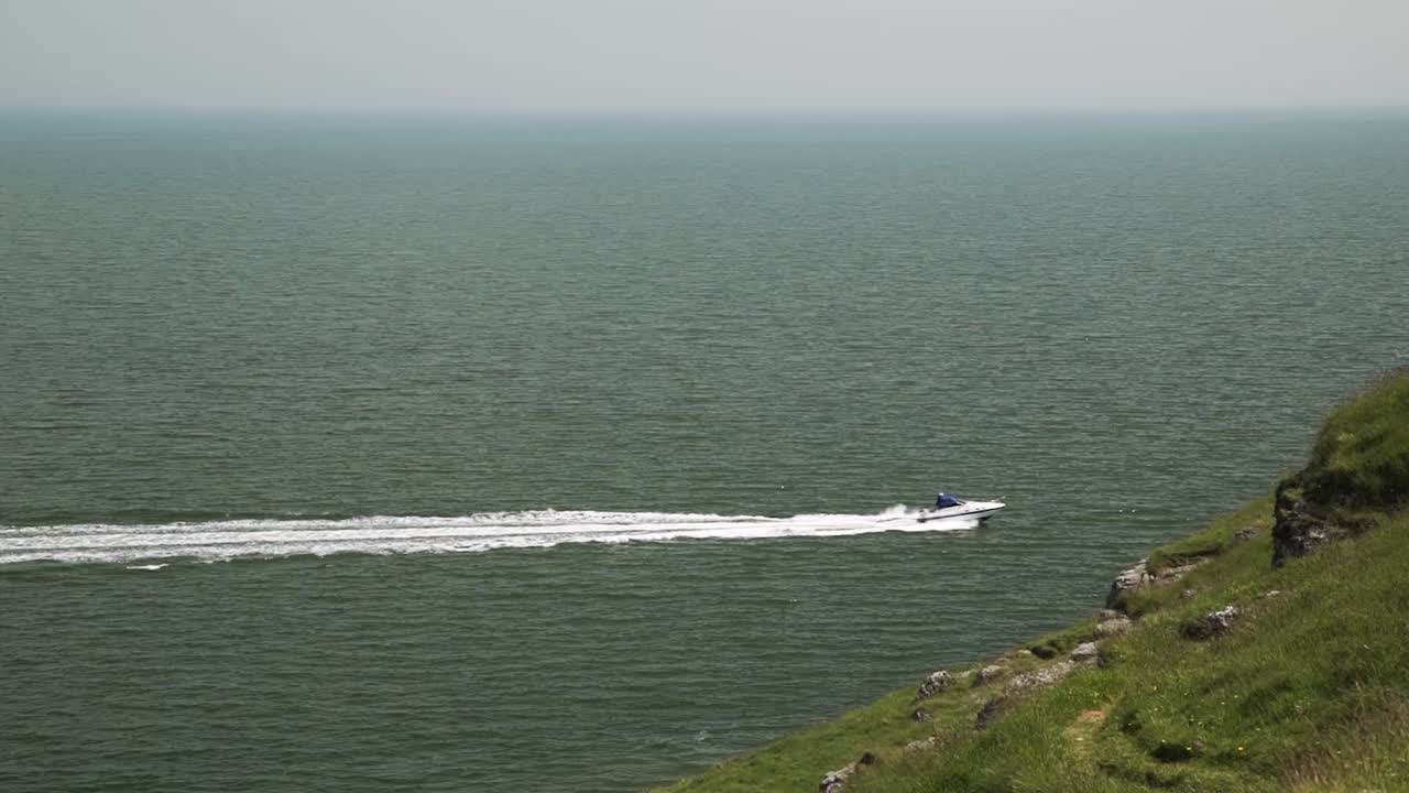 Motorboat speeds across ocean leaving sea trail, disappearing behind grassy cliff edge