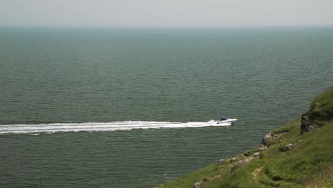 motorboat speeds across ocean leaving sea trail, disappearing behind grassy cliff edge