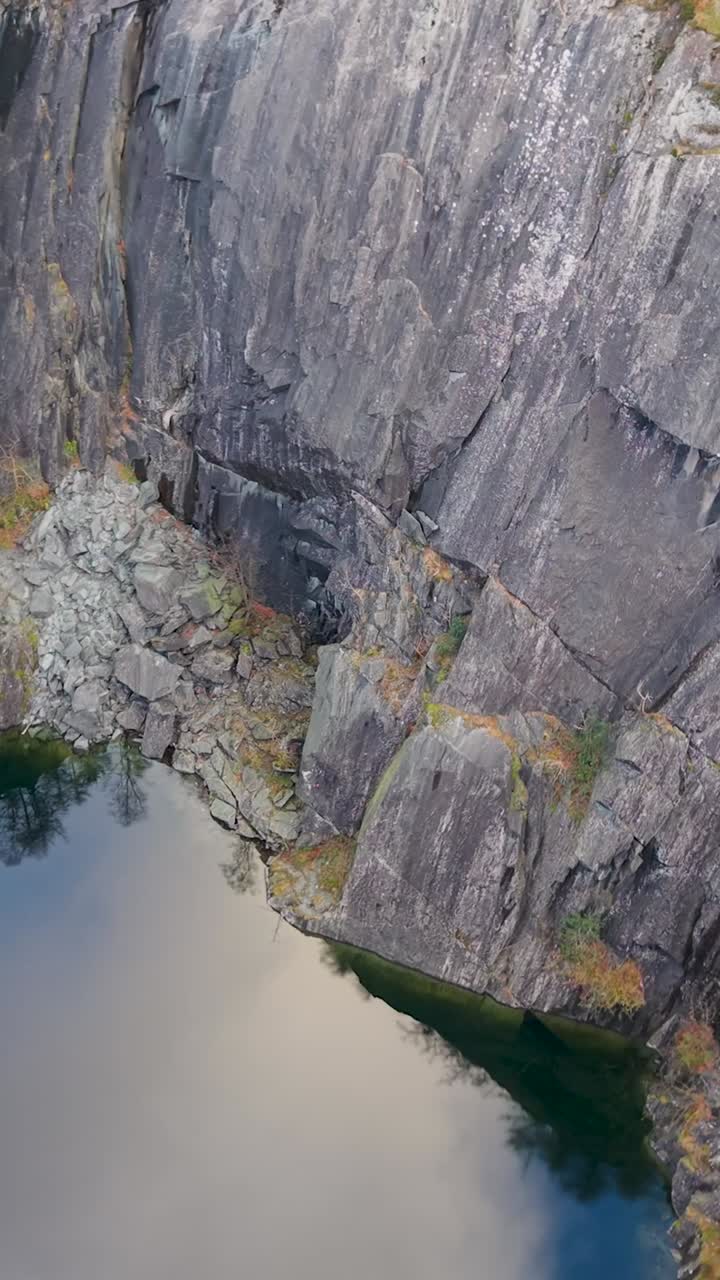 Orbita verticale di un drone sopra la cava e il lago di Hodge Close, Cumbria, che cattura pareti di ardesia, ingresso di una grotta, riflesso delle nuvole nell'acqua e un vibrante fogliame autunnale verde e arancione lungo i bordi della cava