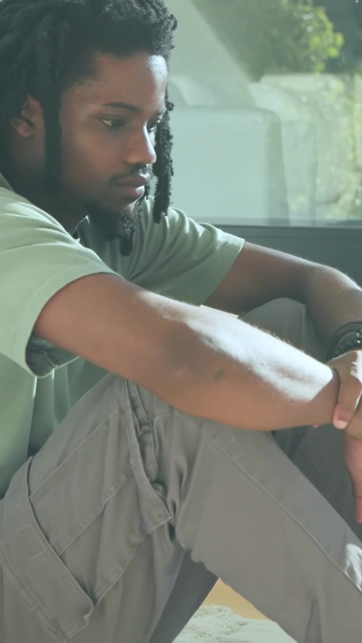Vertical video: Sitting man in green tee adjusting hands, looking at window beside sofa at home