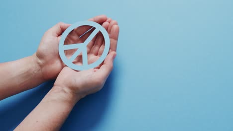 close up of hands of caucasian woman holding blue peace sign with copy space on blue background