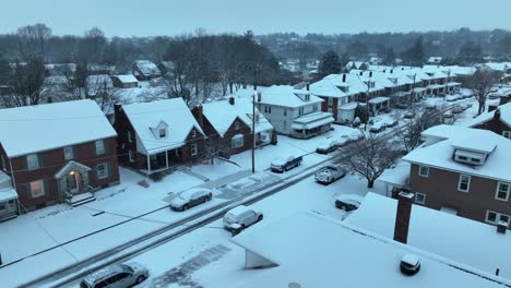 snowy residential area with brick houses and parked cars, trees in the background